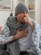 aching ill man with scarf around neck wearing winter hat wrapped in plaid coughing keeping fist close to mouth sitting on couch at living room 1