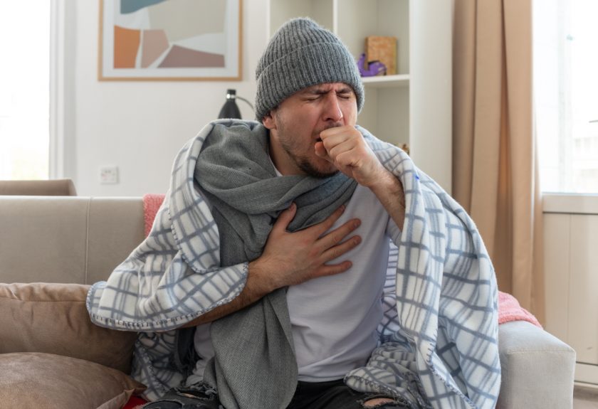 aching ill man with scarf around neck wearing winter hat wrapped in plaid coughing keeping fist close to mouth sitting on couch at living room 1