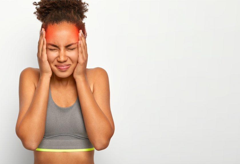 horizontal shot of dissatisfied curly haired woman touches both temples suffers from migraine wears sports bra closes eyes from bad feelings isolated on white wall